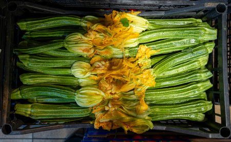 Bright green zucchini and vibrant yellow squash flowers fill a basket in a market setting.の写真素材