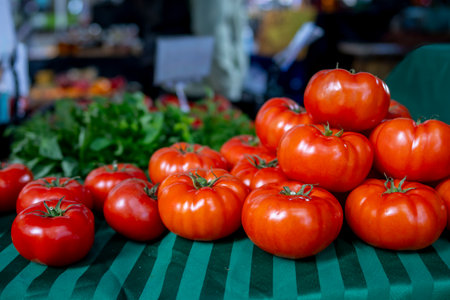 Vibrant red tomatoes are stacked on a green-striped tablecloth at an open-air market. Fresh vegetables surround them, showcasing the variety of seasonal produce available for shoppers.の写真素材