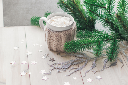 White cup with marshmallows in a warm knitted jacket on a light wooden background with a Christmas tree and silvery stars. space for textの写真素材