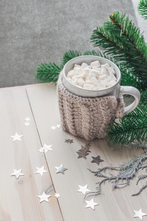 White cup with marshmallows in a warm knitted jacket on a light wooden background with a Christmas tree and silvery stars. space for textの写真素材
