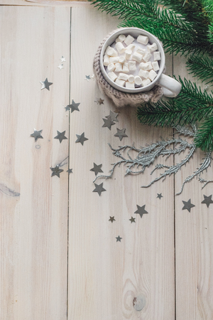 White cup with marshmallows in a warm knitted jacket on a light wooden background with a Christmas tree and silvery stars. space for textの写真素材