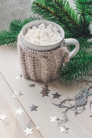 White cup with marshmallows in a warm knitted jacket on a light wooden background with a Christmas tree and silvery stars. space for textの写真素材