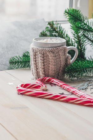 cup in a knitted jacket with two red caramel Christmas candies that lie in the shape of a heart on a white wooden background. Selective focusの写真素材