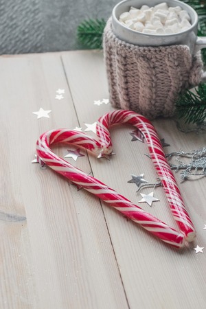 cup in a knitted jacket with two red caramel Christmas candies that lie in the shape of a heart on a white wooden background. Selective focusの写真素材