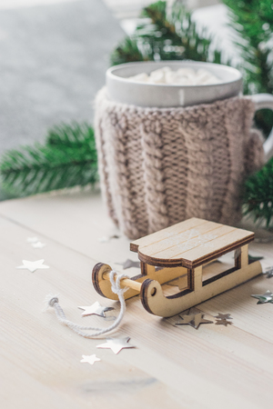 White cup for cocoa in beige knitted clothes. Santa Claus sleigh on a white wooden background with christmas tree space for textの写真素材