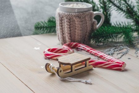 White cup for cocoa in beige knitted clothes. Santa Claus sleigh on a white wooden background with christmas tree and red caramel candy space for textの写真素材