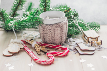 White cup for cocoa in beige knitted clothes. Santa Claus sleigh, mittens, hat, sweater and socks on a white wooden background with christmas tree and red caramel candy space for textの写真素材