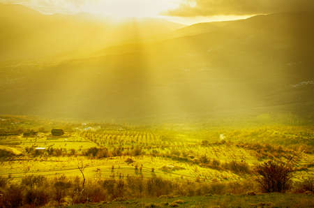 Rural sunset landscape with green hills, dramatic sky, mountains and sun raysの写真素材