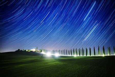 Beautiful Tuscany night landscape with star trails on the sky, cypresses and shining road in green meadow. Natural outdoor amazing fantasy background.の写真素材