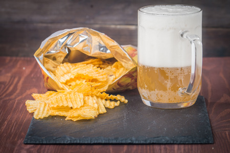Beer with foam in glass mug and potato chips in bag on wooden tableの写真素材