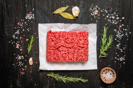Minced meat on paper with seasoning and fresh rosemary on black background, top view