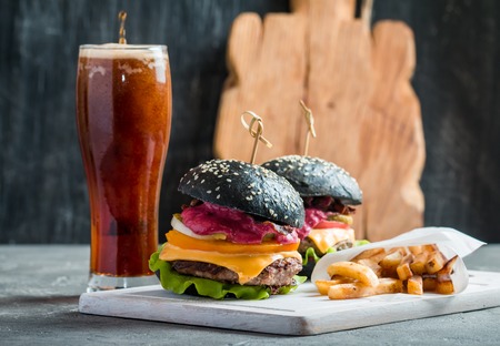 Homemade burger with black bun, beef meat and red berries sauce with glass of dark beer and French fries over wooden table with black background.の写真素材