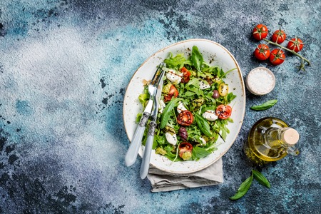Salad with arugula, cherry tomatoes, cream cheese, pesto sauce and herbs on white ceramic plate over rustic blue background, top view, copy spaceの写真素材