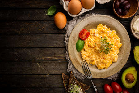 Fresh cooked scrambled eggs in a plate on dark rustic wooden background, top view