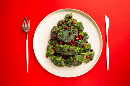 Christmas tree made from broccoli and cranberry on white plate and red background, top view