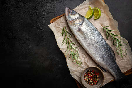 Raw seabass fish with rosemary and spices on dark background, top view