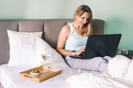 Distance learning online education or work or chating with friends. Happy woman lying in a bed, using laptop and drinking martini.の写真素材