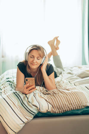 woman enjoy music and looking at smart phone with headphones lying on bed at homeの写真素材