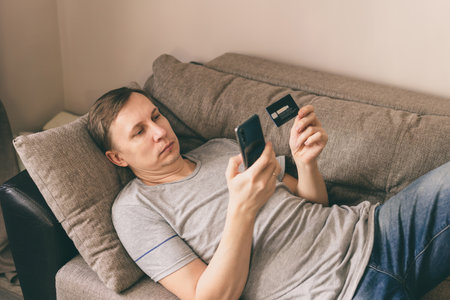 Man holding smart phone and credit card lying down on a couch. Shopping Online at home during lockdown. Vintage or pastel effected photo.の写真素材