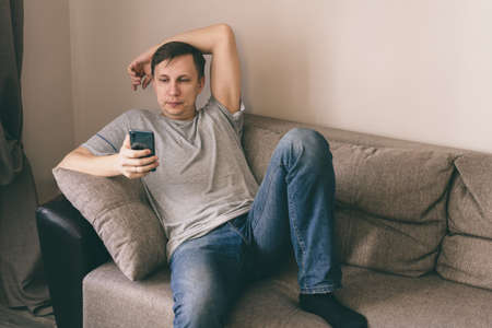Serious man having rest using smartphone sitting on sofa at home
