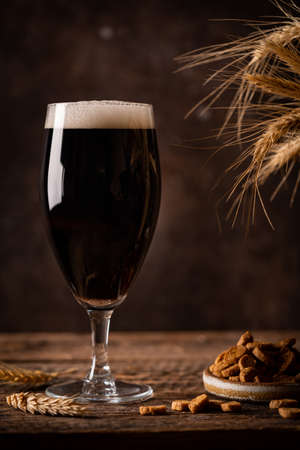 Glass of dark beer with foam head on dark wooden background with empty bottles and beer snacksの写真素材