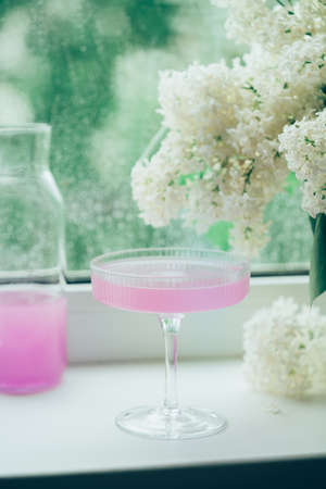 Fresh pink non alcoholic cocktail in glass with lilac flowers against window background.の写真素材