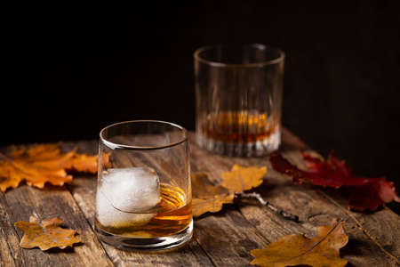 Glass of scotch whiskey and ice on wooden background with autumn leaves
