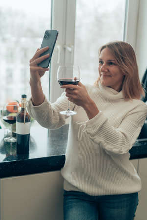 Happy relaxed young woman standing in kitchen with glass of red wine and using her smartphoneの写真素材