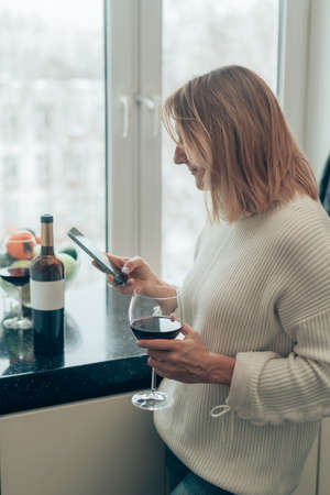 Happy relaxed young woman standing in kitchen with glass of red wine and using her smartphoneの写真素材