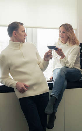 Romantic happy young couple in white clothes holding glasses of wine standing in modern cozy white kitchen, happy married husband and wife drinking red wine celebrate together at homeの写真素材