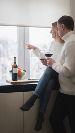 Romantic happy young couple in white clothes holding glasses of wine standing in modern cozy white...