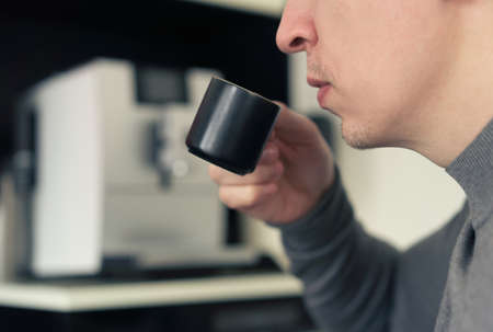 Portrait of Handsome young man drinking coffee espresso in the kitchen at home against coffee machine as a backgroundの写真素材
