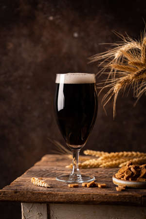Glass of dark beer with foam head on dark wooden background with empty bottles and beer snacksの写真素材