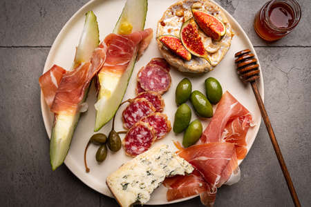 Prosciutto ham, slices of melon, brie and gorgonzola cheese, honey and olives on gray background,...