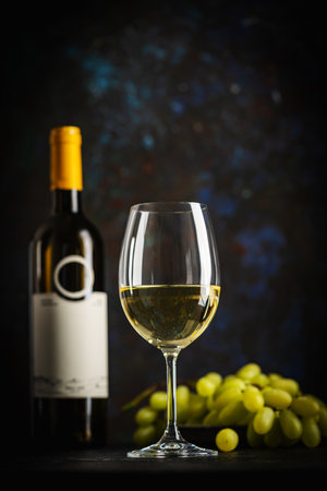 A dark, elegant still life of white wine in a glass with wine bottles and fresh grapes in the background. Perfect for wine-themed compositionsの写真素材