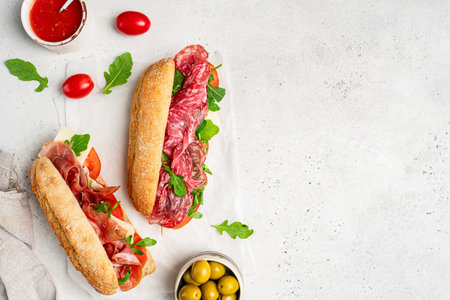 Assorted delicious baguette sandwiches filled with thinly sliced ham or salami and fresh green lettuce or arugula for breakfast arranged on white background, top view with copy spaceの写真素材