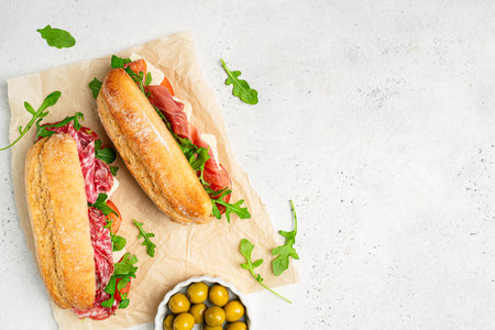Assorted delicious baguette sandwiches filled with thinly sliced ham or salami and fresh green lettuce or arugula for breakfast arranged on white background, top view with copy spaceの写真素材