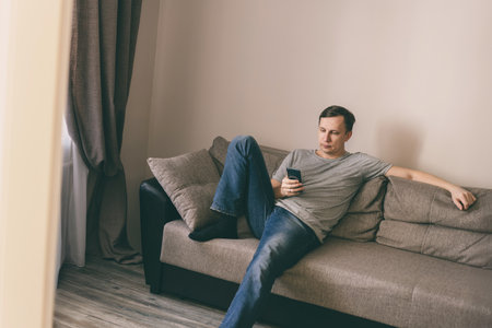 Serious man having rest using smartphone sitting on sofa at homeの写真素材