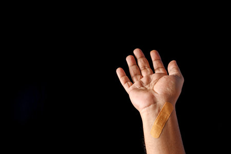 A close-up image of a hand with a small adhesive bandage on the wrist, highlighting themes of care, healing, and injury recovery. The neutral black background enhances the focus on the subject.の写真素材