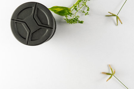 Flat lay composition featuring a black camera lens cap alongside green plants on a white surface, creating a clean and modern aesthetic.の写真素材