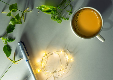 A refreshing composition featuring a white cup of tea surrounded by greenery, glowing light strings, and a pen on a minimalistic background, evoking calmness and creativity.の写真素材