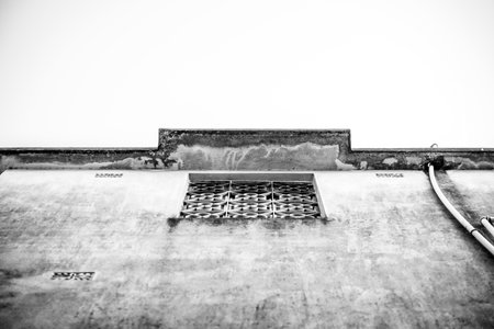 A low-angle view of an aged concrete structure featuring a decorative metal window grill, showcasing textures and architectural decay in dramatic black-and-white tones, evoking a stark and minimalistic aesthetic.の写真素材