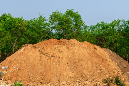 A large mound of brown soil is set against a backdrop of lush green trees under an expansive blue sky, creating a contrast of earthy and natural beauty.の写真素材