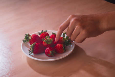 a hand picking a strawberry from a white plate with red strawberriesの写真素材