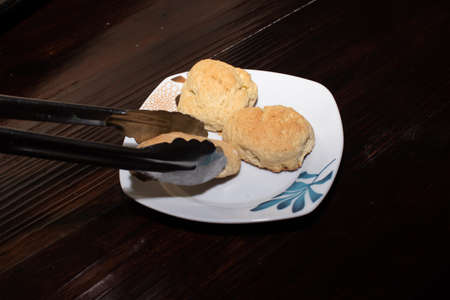 some metal tongs taking a piece of bread, from a white plate with three pieces of homemade bread, on a wooden tableの写真素材