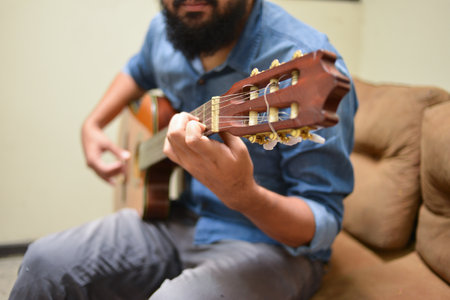 Man dressed in casual clothes playing guitar, sitting on an armchair in a roomの写真素材