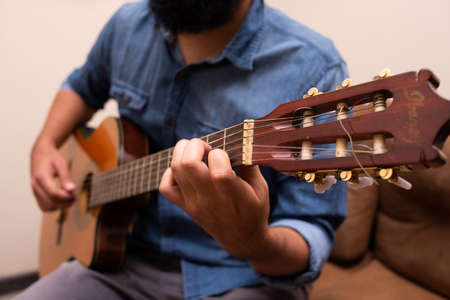 Man dressed in casual clothes playing guitar, sitting on an armchair in a room