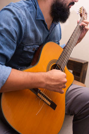 Man dressed in casual clothes playing guitar, sitting on an armchair in a room