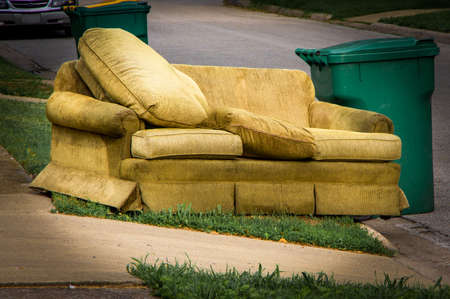 Old yellow couch with cushions sits on a curb in residential area next to a recycling cart waiting for bulk waste hauler pickup on trash dayの写真素材