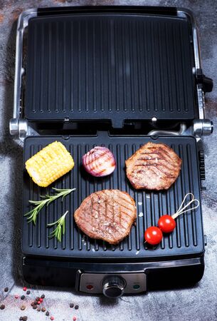 Preparation beef steaks and vegetables on an electric grill on a gray concrete background, top viewの写真素材